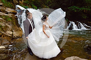 Wedding, couple near beautiful grand waterfall in mountain. Wind fluttering a long veil. Landscape of hills and mountains