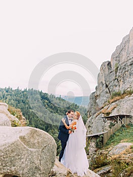 Wedding couple standing at rocky mountains against the sky and embracing. Cute romantic moment.