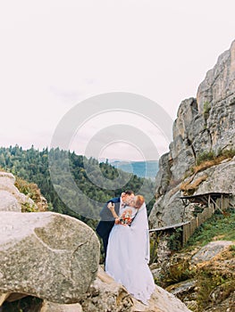 Wedding couple standing at rocky mountains against the sky and kissing. Cute romantic moment.