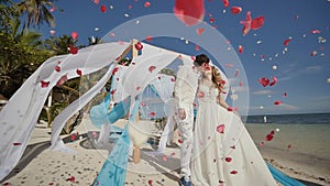 A wedding couple on a tropical beach next to the ocean. Kiss under the arch with white and blue air wings flying in the