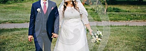 Wedding couple walking in the green park. Curvy bride in white lace dress and groom are holding hands. Overweight happy people.