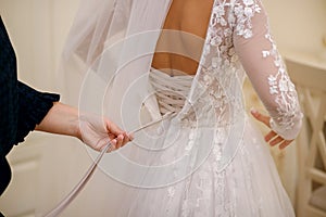 On the wedding day, the bride wears a white wedding dress. A faithful friend helps to tie ribbons on a corset, back view