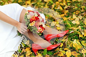 Wedding day. Close up of bride`s feet in red shoes, colorful bridal bouquet and autumn leaves on green grass