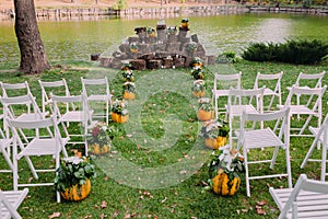 Wedding decoration with autumn pumpkins and flowers. Ceremony outdoor in the park. White chairs for guests