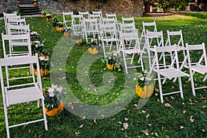 Wedding decoration with autumn pumpkins and flowers. Ceremony outdoor in the park. White chairs for guests