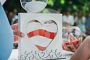 Wedding decorative heart for ceremony. Red and white sand in the glass and wooden form. bride and groom hands with golden rings.