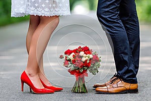 Wedding details: stylish red and brown shoes of bride and groom. Bouquet of roses standing on the ground between them. Newlyweds s