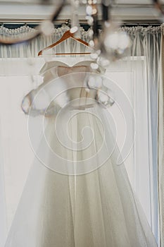 Wedding dress. White wedding dress with a full skirt on a hanger in the room of the bride with white curtains. Wedding