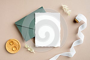 Wedding invitation card mockup, green envelope, golden rings, silk ribbon, dried flowers on pastel beige background. Flat lay, top