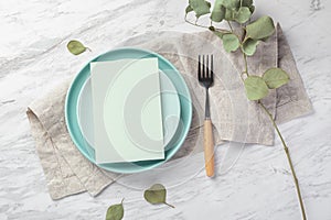 Wedding menu card mockup on plate with linen napkin on marble table with green leaves