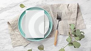 Wedding menu card on plate with fork on linen napkin on marble table with green leaves