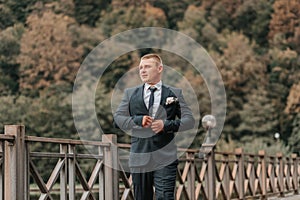 Wedding portrait. A man in a dark gray suit zips up his jacket as he walks across the bridge and looks away.