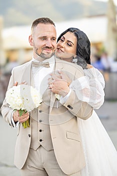 Wedding portrait of smiling newlyweds. A stylish groom in a beige suit and a cute brunette bride in a white dress are tenderly