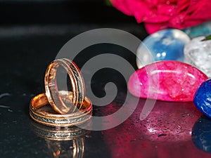 Wedding rings in focus on a dark reflective surface and colorful gem stone out of focus in the background. Concept romance, love