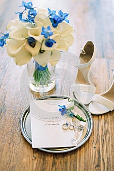 Wedding rings lie on a round tray next to the invitation near a wedding bouquet of flowers in a vase