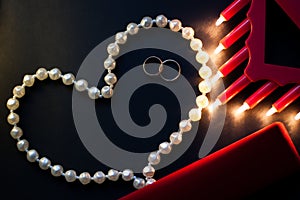 Wedding rings, a string of pearls on a black background, burning lights, a red box. St. Valentine`s Day