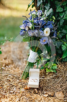 Wedding rings in a white box on dry grass with a bride`s wedding bouquet of blue flowers.