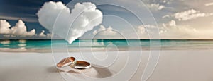 Wedding rings on white sandy beach with a heart shaped cloud in the sky. Panorama with copy space.