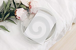 Wedding still life scene. White empty book cover mockup on marble tray. Pink peony flowers on white linen table cloth