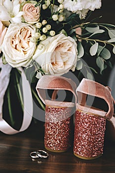 Wedding velvet pink shoes with shiny beautiful heels with gold wedding rings beside a bouquet of white roses, eucalyptus on a dark