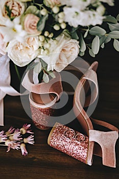 Wedding velvet pink shoes with shiny beautiful heels with gold wedding rings beside a bouquet of white roses, eucalyptus on a dark