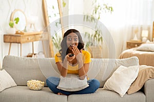 Weekend entertainments. Shocked African American woman with TV controller enjoying interesting movie indoors