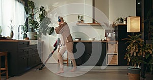 Weekend fun. Happy cheerful 60s senior woman dancing to music in headphones, cleaning floor with cordless vacuum cleaner