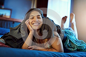 Weekends in this house are clothing optional. Portrait of a happy young woman lying under a pile of clothes on her bed.