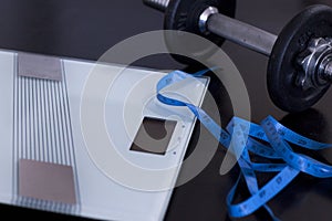 Weighing scale and  measuring tape with dumbbell on the side on black table background.