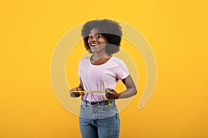 Weight loss concept. Fit african american lady measuring her waist with tape over yellow studio background