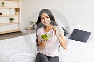Weight loss, slimming diet concept. Happy Indian woman eating yummy vegetable salad on bed at home