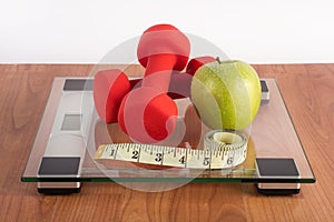 Weight scale with red dumbbell, measuring tape and fresh green apple on wooden floor