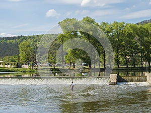 Weir on the river Berounka in village Zadni Treban with fly fisherman standing in the stream and lush green trees. Sunny