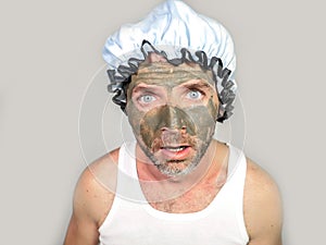 Weird looking man with shower cap and cream on his face horrified seeing himself ugly on bathroom mirror applying facial mask male