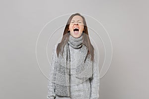 Weird young woman in gray sweater, scarf keeping eyes closed, mouth wide open screaming on grey background
