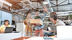 Welcome on board. Young man in casual wear holding box with things and giving high five to his new coworker while