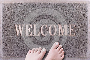 Welcome Door Mat With Female Feet. Friendly Grey Door Mat Closeup with Bare Woman Feet Standing. Welcome Carpet.