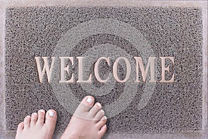 Welcome Door Mat With Female Feet. Friendly Grey Door Mat Closeup with Bare Woman Feet Standing. Welcome Carpet.
