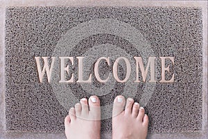 Welcome Door Mat With Female Feet. Friendly Grey Door Mat Closeup with Bare Woman Feet Standing. Welcome Carpet.