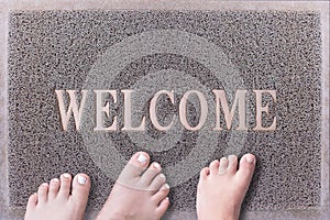 Welcome Door Mat With Three Feet. Friendly Grey Door Mat Closeup with Bare Feet Standing. Welcome Carpet.