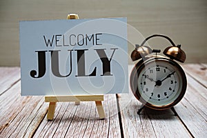 Welcome July text with alarm clock on wooden background