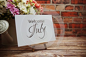 Welcome July text with flower bouquet decoration on wooden and old brick wall background