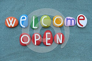 Welcome Open, creative logo composed with multi colored stone letters over green sand