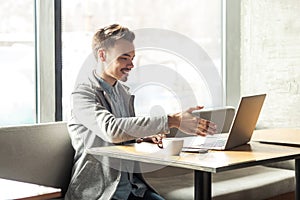 Welcome! Portrait of satisfied happy young man in grey blazer are sitting in cafe and greeting a worker through a webcam and