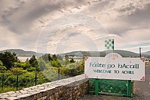 Welcome to Achill sign in Irish and English language by road. Travel and tourism information. Cloudy sky. Small county colors flag