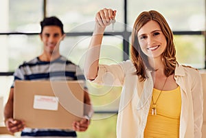 Welcome to our new home. Portrait of a young woman holding up the keys to a new home with her boyfriend carrying boxes