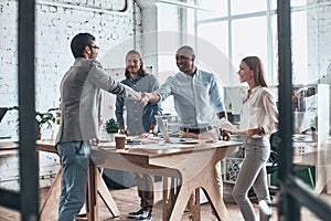 Welcome to our team. Young modern men in smart casual wear shaking hands while standing with their colleagues in the board room