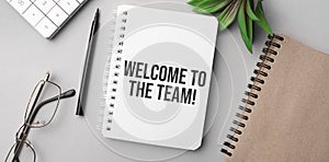 welcome to the team is written in a white notebook with calculator, craft colored notepad, plant, black marker and glasses