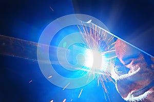 Welder in a protective mask in a dark shop floor weld metal parts. By welding sparks fly in different directions