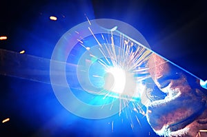 Welder in a protective mask in a dark shop floor weld metal parts. By welding sparks fly in different directions